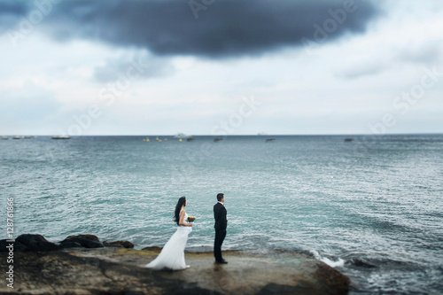 Bride stands behind the groom on the rocks over calm grey sea