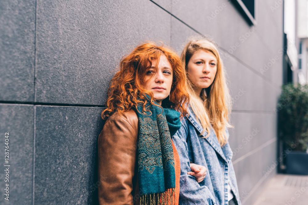 Two young handsome caucasian blonde and redhead woman friends posing outdoor in the city - friendship, interaction concept Two young handsome caucasian blonde and redhead woman friends posing outdoor in the city - friendship, interaction concept