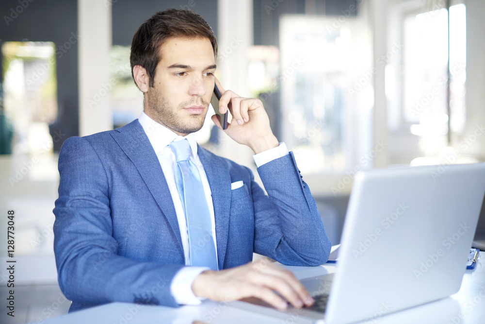 Financial professional man in the office. Shot of a young businessman making call and while sitting at desk in front of laptop using internet banking. Financial professional man in the office. Shot of a young businessman making call and while sitting at desk in front of laptop using internet banking.