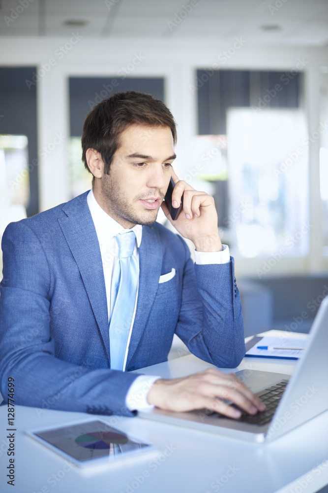 Successful professional man. Shot of a young confident businessman making call while sitting at desk in front of laptop and working online. Successful professional man. Shot of a young confident businessman making call while sitting at desk in front of laptop and working online.