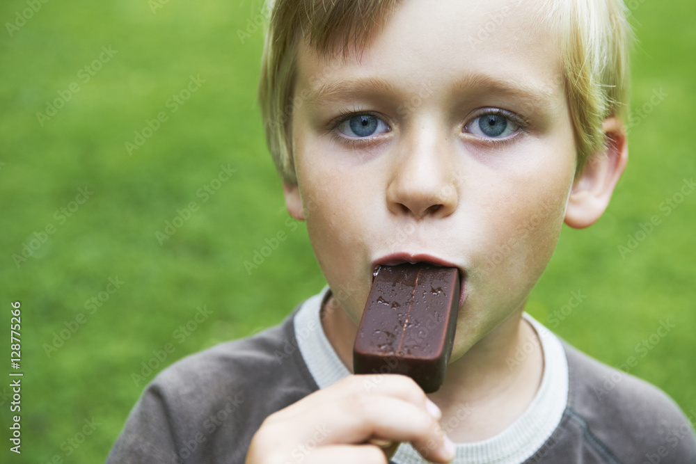 Portrait of young child blond boy eating a tasty ice cream outside in the garden with green grass background.
Portrait of young child blond boy eating a tasty ice cream outside in the garden with green grass background.