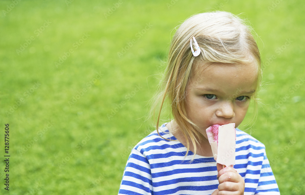Beautiful little girl eats ice-cream in the summer with green grass background.
Beautiful little girl eats ice-cream in the summer with green grass background.