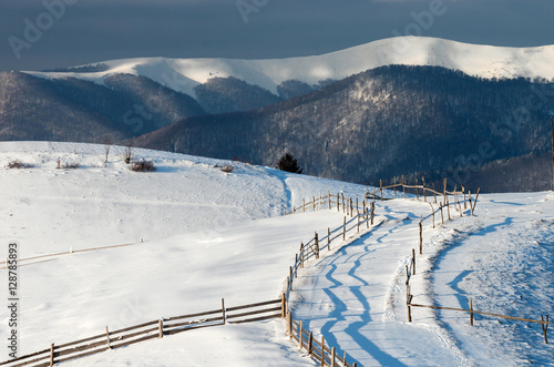 Pasture fence on snowy hills in the sunlight against the background of a mountain range and a dark sky. A fabulous winter landscape.