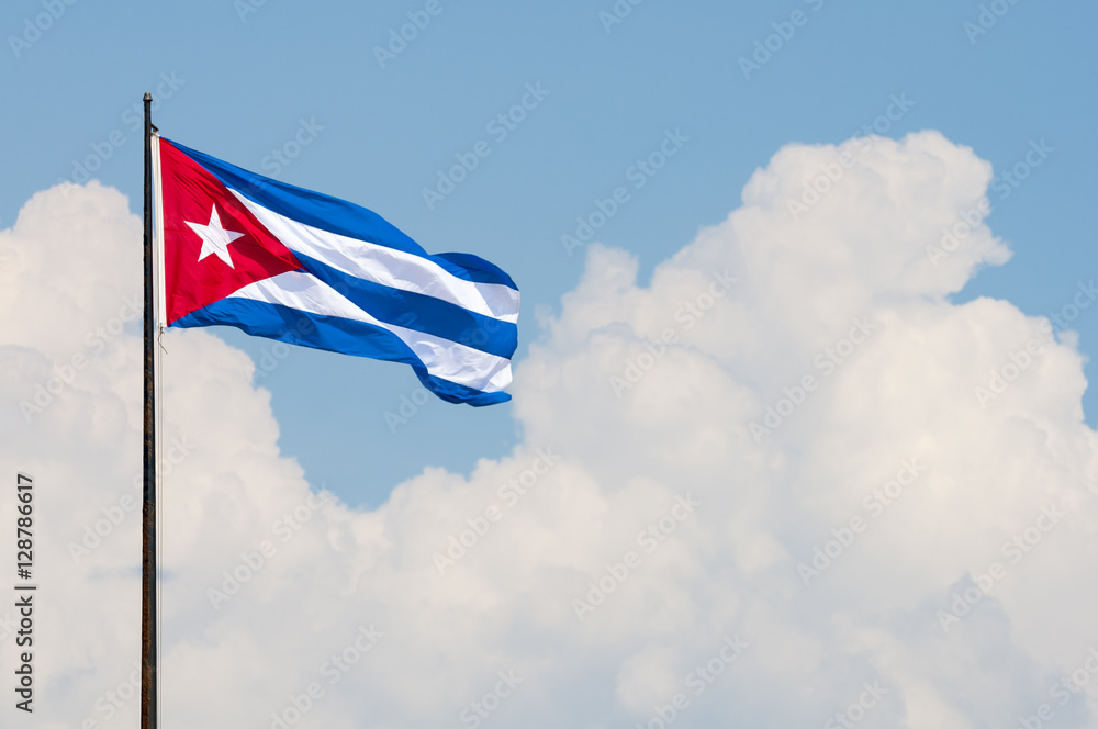 Cuban flag flying from a flagpole in the wind in front of a soft blue sky with tropical clouds in Havana, Cuba