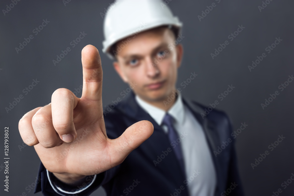 Handsome businessman in a helmet standing on gray background he presses the finger, focus on hand