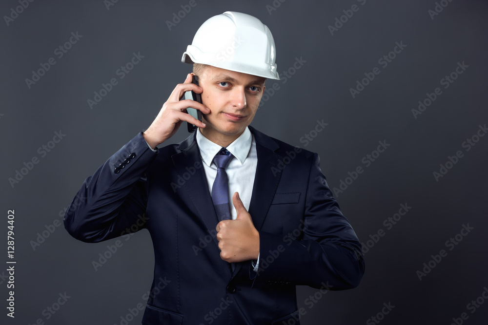 Handsome young engineer in a helmet talking on the phone and showing thumbs up standing on gray background Handsome young engineer in a helmet talking on the phone and showing thumbs up standing on gray background