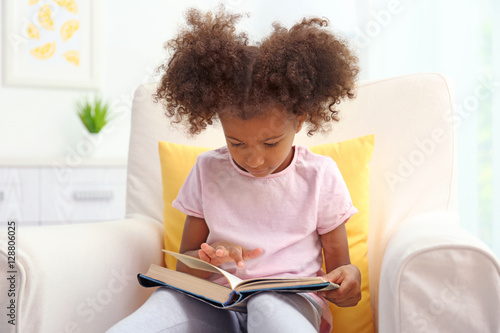 Little African American girl sitting in armchair and reading book in room