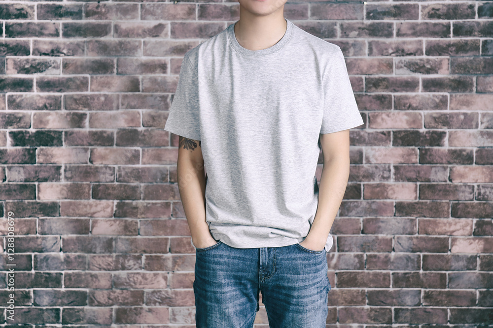 Young man in blank grey t-shirt standing against brick wall, close up Young man in blank grey t-shirt standing against brick wall, close up