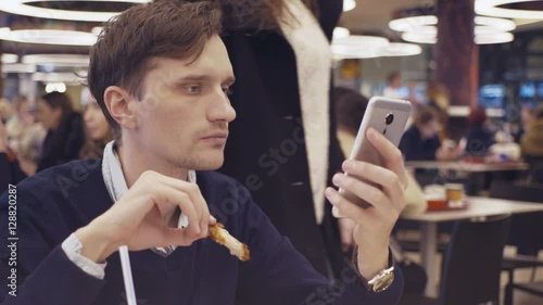 Young men using smartphone eat fried chicken wings at food court in shopping mall. Portrait