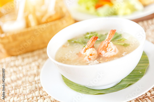 Traditional Thai porridge rice gruel and shrimp in white bowl with seasoning background on wood table in sunlight.