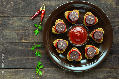 Sandwiches with black rye bread in the shape of a heart, blood sausage (Morcillo) and pieces of sweet pepper on skewers in a ceramic bowl with tomato sauce on a dark wooden background. The top view. photo