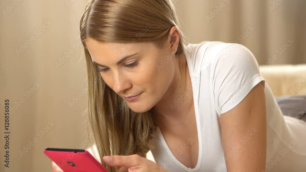 Beautiful attractive young woman in a white t-shirt with a pink smartphone lying on a sofa. Browsing Internet and smiling. Beautiful attractive young woman in a white t-shirt with a pink smartphone lying on a sofa. Browsing Internet and smiling.