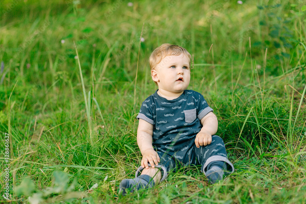 chubby little boy in dark clothes sitting on the grass in the park on a summer day chubby little boy in dark clothes sitting on the grass in the park on a summer day