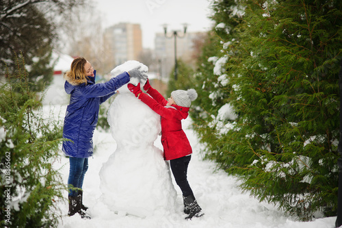 Mother with daughter of younger school age build a snowman in the park.