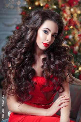 Christmas portrait of a beautiful woman with chic curls in a red dress on a background of the Christmas tree.