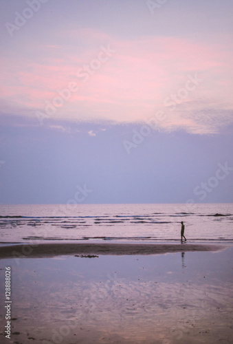 Silhouette of a young man walking along the shoreline of a tranquil beach at sunset at dusk with reflection of the sea and clouds  Goa  India