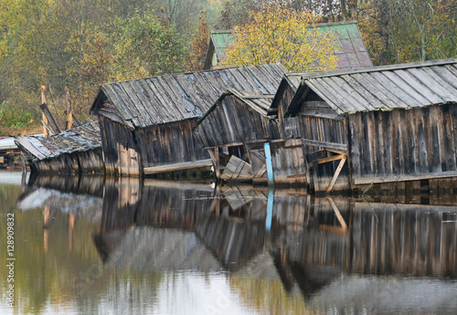 Old dilapidated barns for boats reflected in river. © Belozorova Elena