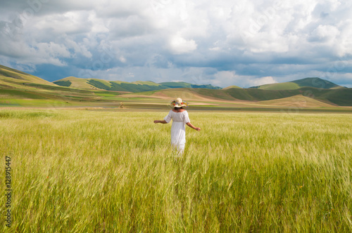 Girl in White dress and straw hat walking among fields in countryside 