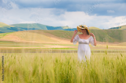 Girl in White dress and straw hat walking among fields in countryside
