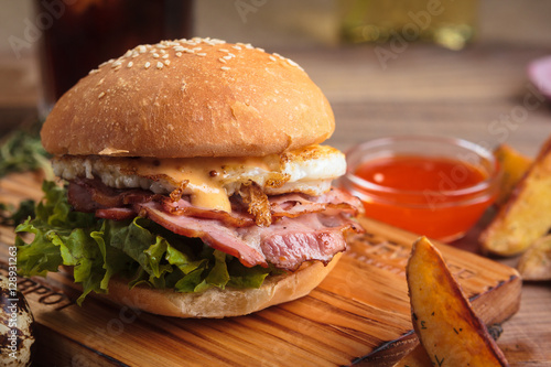 Concept: restaurant menus, healthy eating, homemade, gourmands, gluttony. Trendy breakfast burger with ham and eggs with ingredients, drinks and potato wedges on messy vintage wooden background.