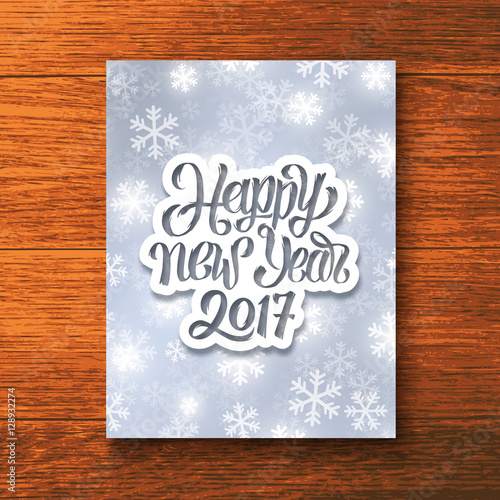Happy New Year 2017 hand lettering text on paper label over glowing winter background. Vector greeting card template with typography for holidays above wooden backdrop.