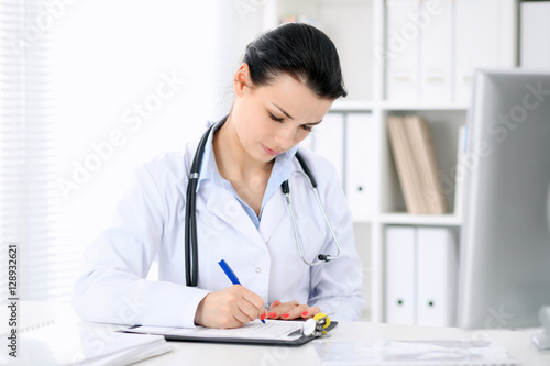 Young brunette female doctor sitting with clipboard near window in hospital and filling up medical history form.