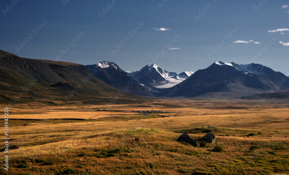A highland river valley with yellow grass on a background of snow covered high mountains and glaciers under clouds and blue sky, Plateau Ukok, Altai Siberia, Russia