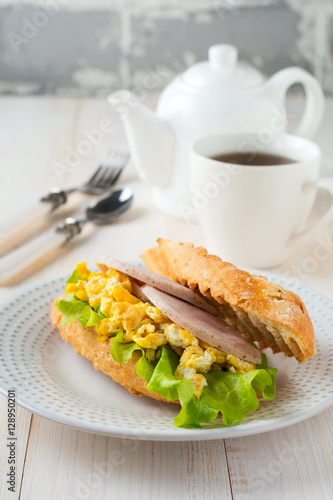 Sandwich stuffed with scrambled eggs, ham and lettuce leaves on a light wooden background. Selective focus.