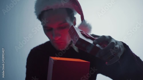 Young man in red Santa Claus hat openinng gift box surprised and happy with what you see photo