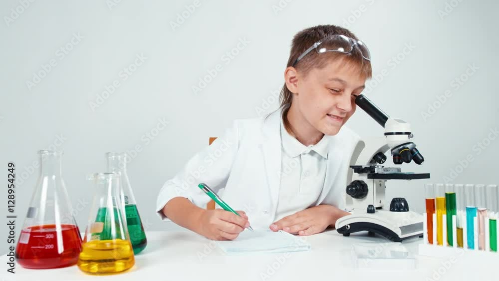 Young chemist looking at microscope and something writing in her notebook and smiling at camera. Child isolated Young chemist looking at microscope and something writing in her notebook and smiling at camera. Child isolated