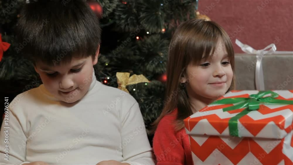 Little kids looking at their christmas gifts near the christmas tree. Little brown-haired girl holding big striped gift box in her hads. Little children showing their christmas gifts to each other Little kids looking at their christmas gifts near the christmas tree. Little brown-haired girl holding big striped gift box in her hads. Little children showing their christmas gifts to each other