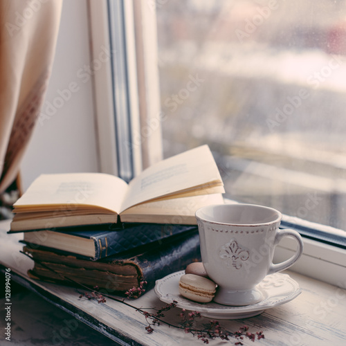 Cozy winter still life: cup of hot coffee and opened book on vintage windowsill against snow landscape from outside