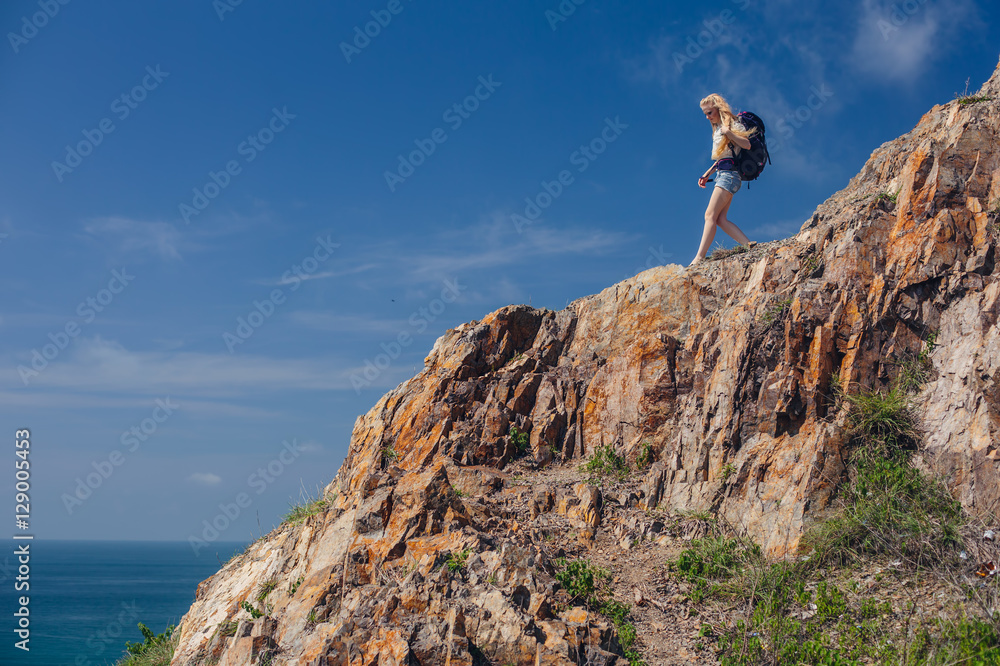 Young woman walking with with backpack on cliff edge to blue ocean valley