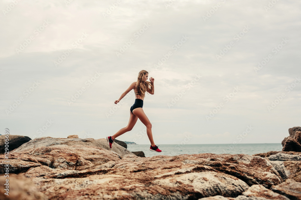 Woman runner training for marathon. Female runner in sporty top jogging on beach rocks