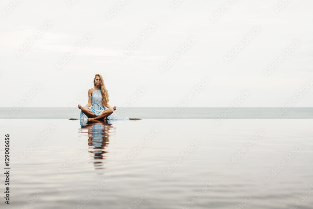 Obraz premium Beautiful female in blue dress sitting near sea beach in lotus pose, relax and meditate. Water Reflection in pool, woman practice