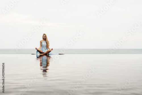 Beautiful female in blue dress sitting near sea beach in lotus pose, relax and meditate. Water Reflection in pool, woman practice