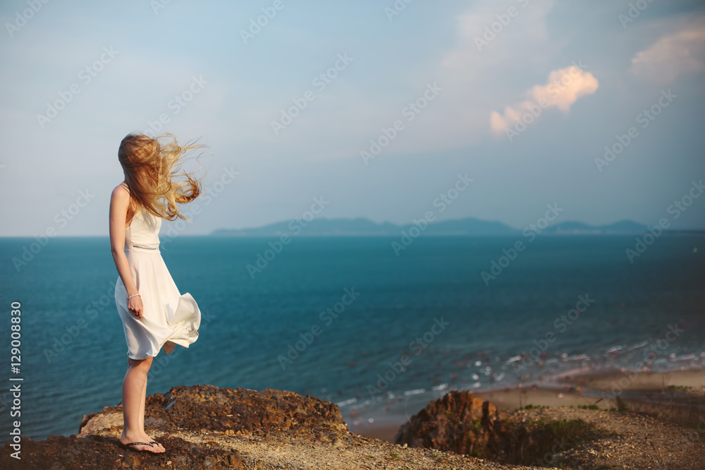 Young girl on rock beach, wind in blond hair and white dress