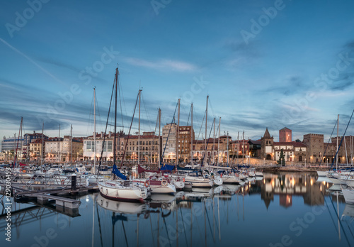 Yatchs and pier in leisure port on maritime fishing district of Gijon, Spain, Europe. Beautiful reflection on calm sea water of boats, buildings, sky at dusk at touristic cultural travel destination. © Starstuff