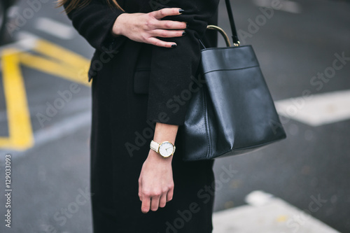 detail of a fashionable woman wearing a black oversized coat, a white and golden watch and a black trendy handbag. perfect fall fashion outfit.