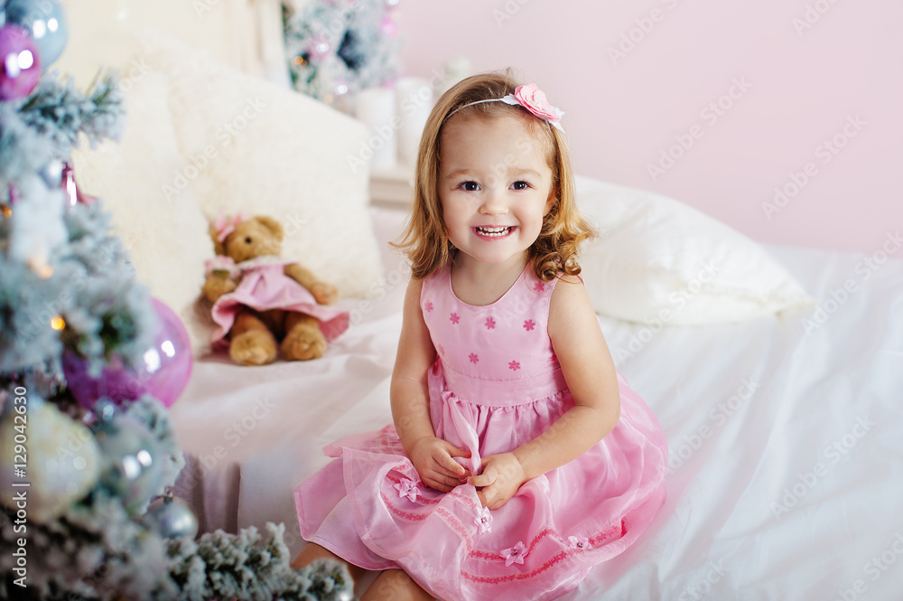 Very nice charming little girl blonde in pink dress sitting on a child's bed and laughs loudly the background of Christmas trees in bright interior the house Very nice charming little girl blonde in pink dress sitting on a child's bed and laughs loudly the background of Christmas trees in bright interior the house
