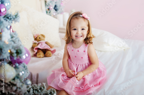 Very nice charming little girl blonde in pink dress sitting on a child's bed and laughs loudly the background of Christmas trees in bright interior the house