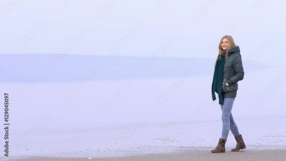 Young beautiful girl walking on sandy beach and smiling. Quiet weather, calm water, fog, mist and haze over water. Beach is reflected on water surface. Fantastic landscape film or movie. Close shot Young beautiful girl walking on sandy beach and smiling. Quiet weather, calm water, fog, mist and haze over water. Beach is reflected on water surface. Fantastic landscape film or movie. Close shot
