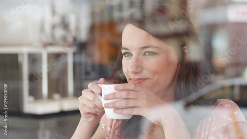 Young woman looking outside through the window in a coffee shop.  She drinks from a cup. She looks happy. We see traffic and people in the reflection of the window.