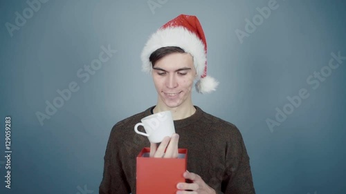Young man in Santa hat opening a gift box with a surprise inside seeing coffee cup photo