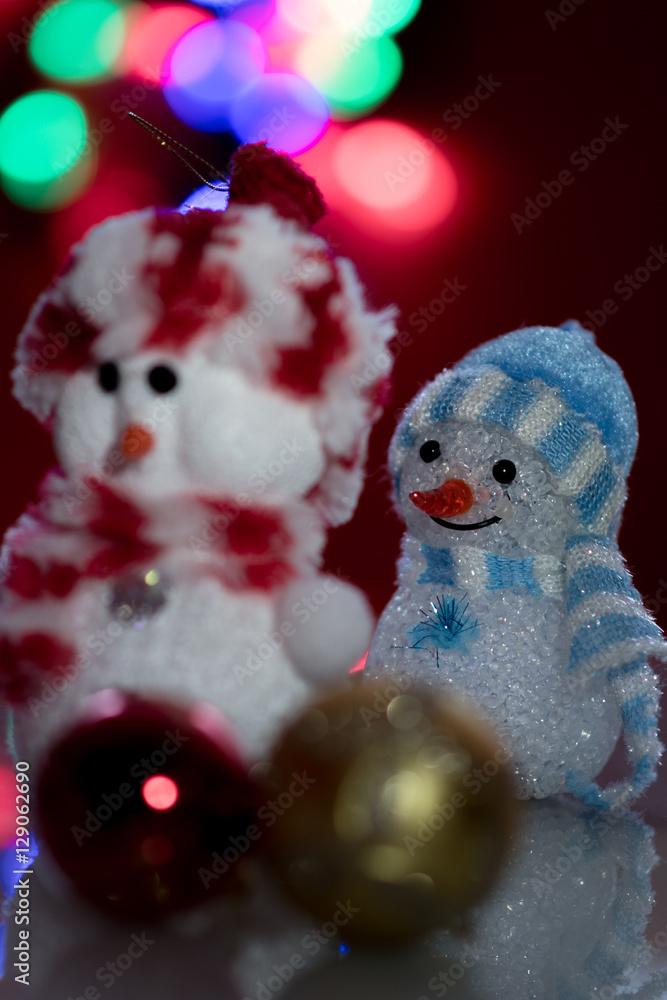 Illuminated two Snowman dolls in front of Christmas tree lights, blurred background. Festive snowman with Christmas light background. Focused on a blue snowman.