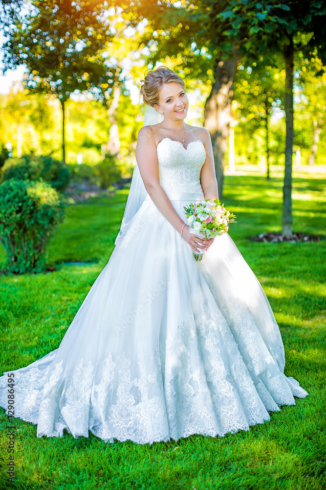 beautiful charming bride in white wedding dress holding bouquet on background of green park