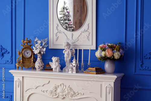Белый комод, на котором часы, вазы с цветами, скульптура и зеркало в котором отражается новогодняя елка

