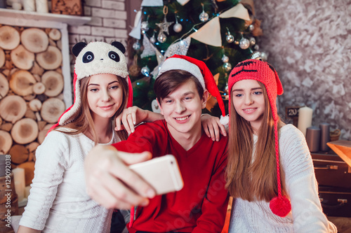 Beautiful girls and boy in funny hats sitting on the floor near a Christmas tree with gifts. Make selfie. New Year.