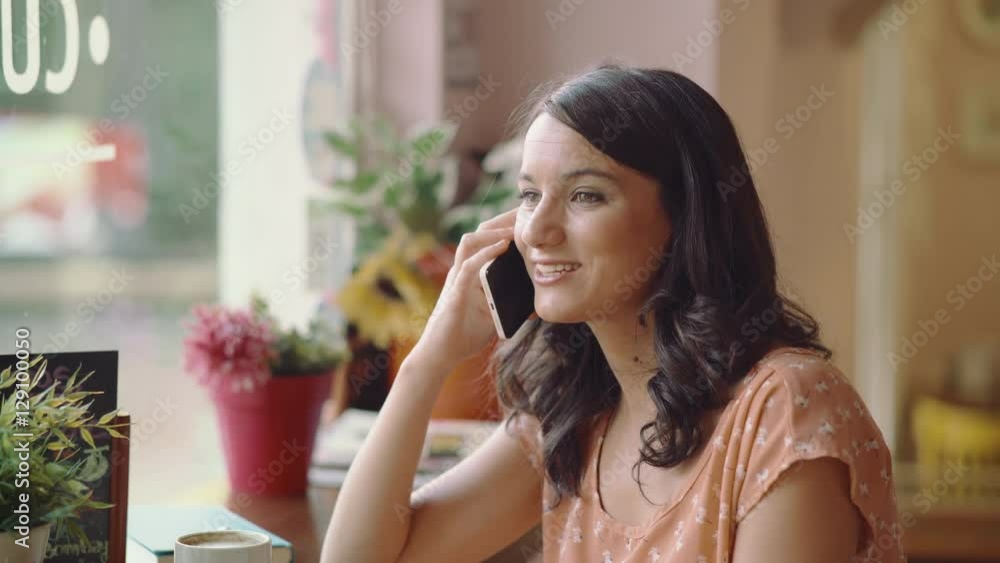 Young woman talks with her smartphone next to the window in a coffee shop. She smiles while talking. Dolly shot. Young woman talks with her smartphone next to the window in a coffee shop. She smiles while talking. Dolly shot.
