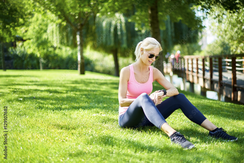 Starting morning outdoor. Full length shot of a fit woman sitting in the grass and checking her sport watch after morning workout. Starting morning outdoor. Full length shot of a fit woman sitting in the grass and checking her sport watch after morning workout.